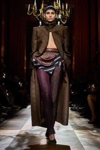 A model wearing a funnel-neck jacquard coat, skinny trousers, and a scarf-like belt while walking at the F/W 25 Dries Van Noten show during Paris Fashion Week.