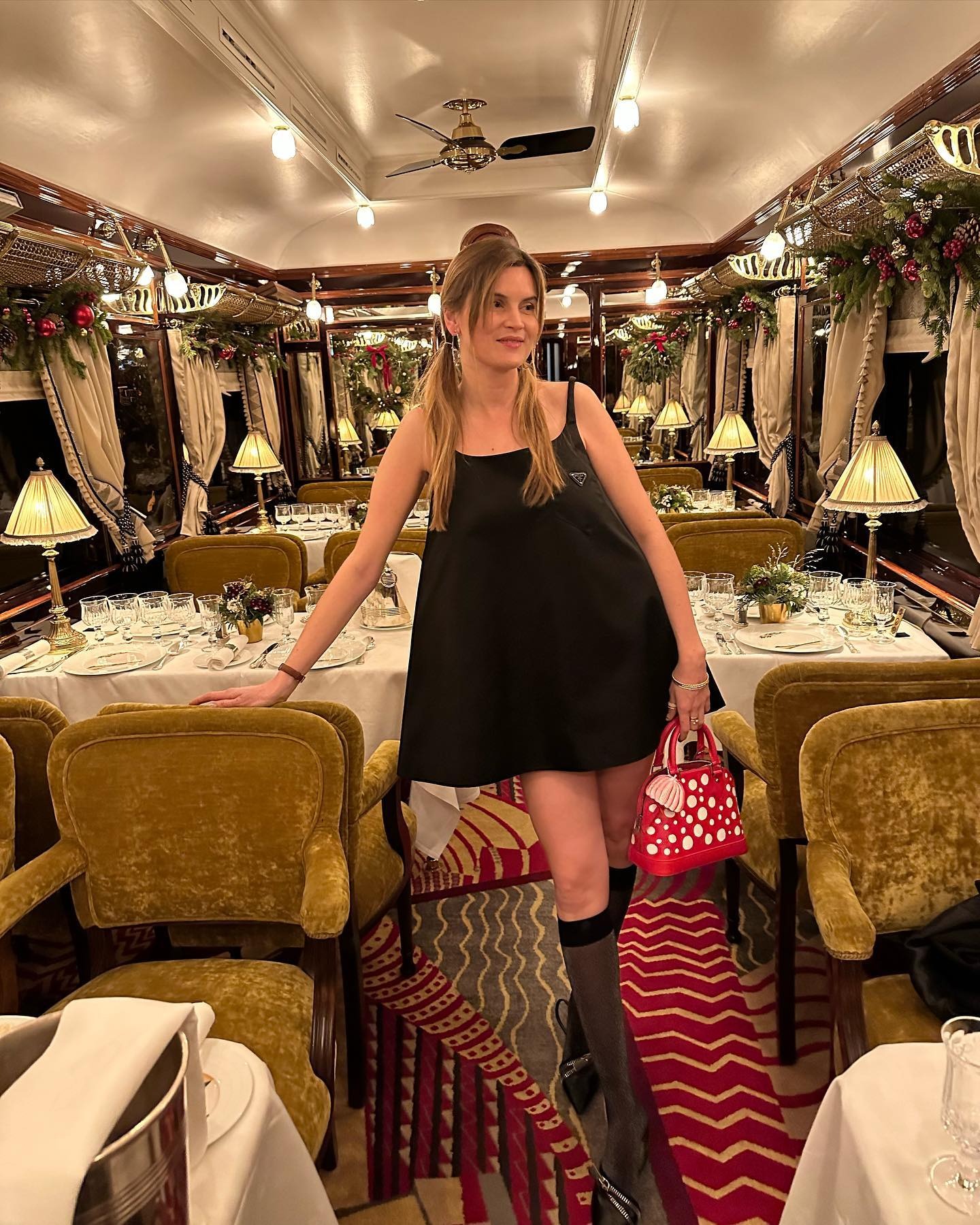 writer, editor, and fashion creative Euge&#769;nie Trochu poses on the Christmas Orient Express Paris/Courchevel wearing a black Para mini dress, a white and red Louis Vuitton x Yayoi Kusama Alma bag, knee-high socks and heels